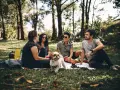 Un gruppo di amici fa un picnic nel parco, un cane bianco e marrone si avvicina. Uno dei ragazzi, sicuro di sé, ride con gli altri.