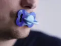 Un uomo con barba corta ha un ciuccio blu in bocca, un'immagine che evoca adulti che si comportano da bambini.