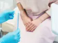 Mani con guanti blu tengono uno strumento per il pap test. La paziente, in attesa, è seduta.
