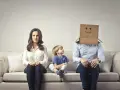 Famiglia seduta sul divano. La madre è triste, il bambino guarda il padre con la testa coperta da un cartone con un sorriso disegnato. Sembra che mio marito stia con me solo per i figli.