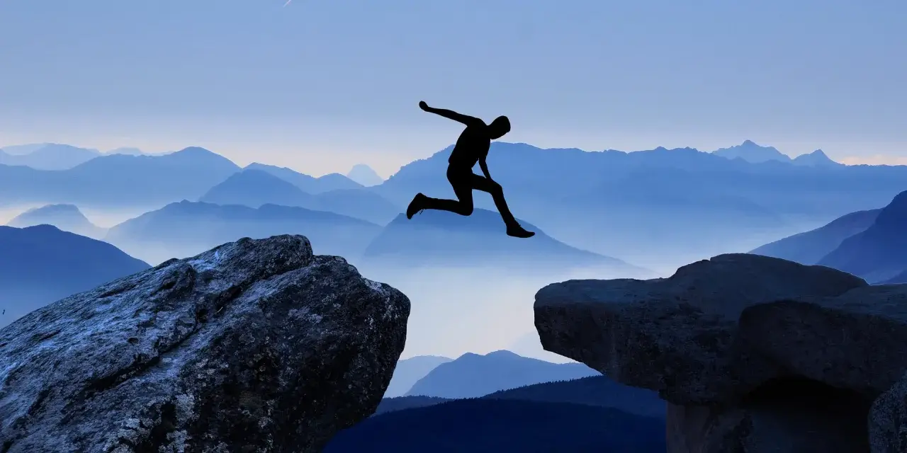 Silhouette di una persona che salta tra due rocce, con montagne blu sullo sfondo. Un viaggio per perdersi per ritrovarsi, un salto nel vuoto per una coppia.