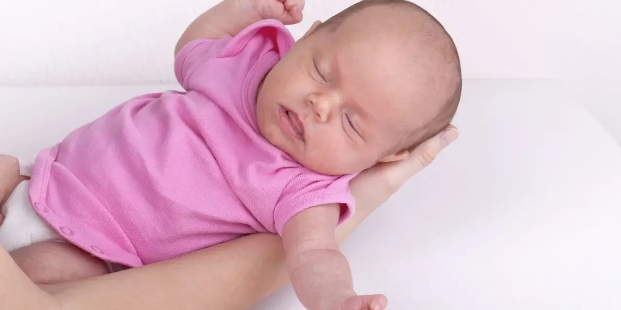 Neonato in rosa si irrigidisce e stringe i pugni, dormendo sereno tra le braccia.
