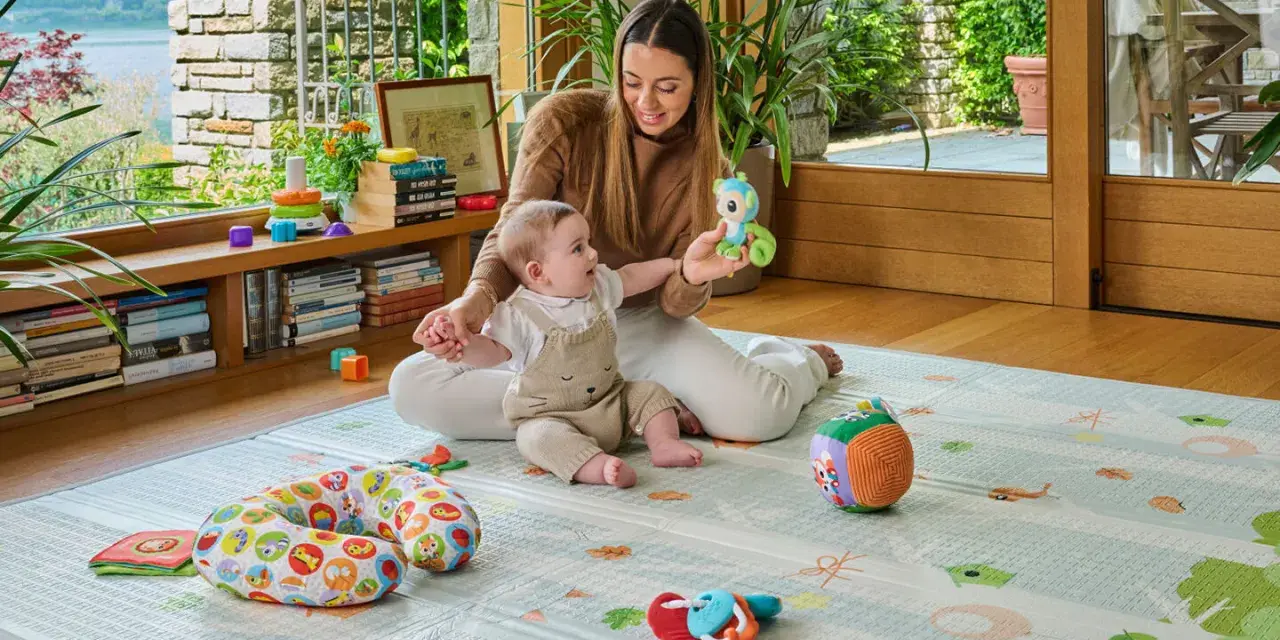 Mamma gioca con un neonato di 6 mesi che non sta seduto, su un tappeto gioco con tanti giocattoli colorati.