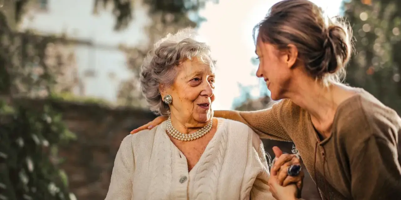 Una donna anziana e una giovane donna si guardano con affetto. La giovane donna ha una mano sulla spalla dell'anziana, un gesto che suggerisce cura e supporto, forse per superare le conseguenze di un cattivo rapporto con la madre.