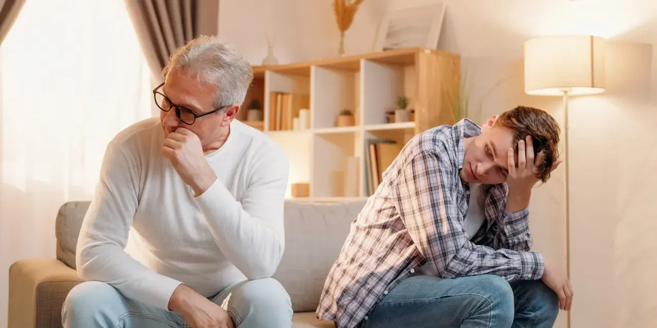 Padre e figlio seduti sul divano, distanti e pensierosi. Crescere in una famiglia dove si litiga sempre lascia segni profondi.