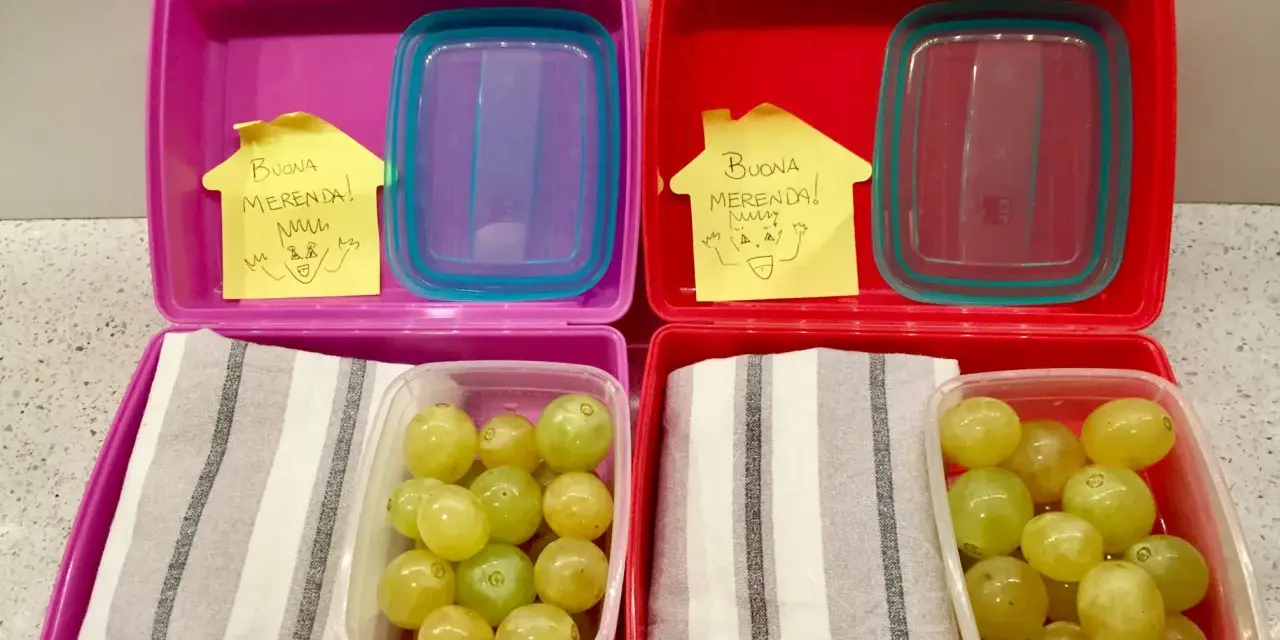 Due lunchbox rosa e rosse pronte per le merende per bambini, con uva e un bigliettino "Buona Merenda!".