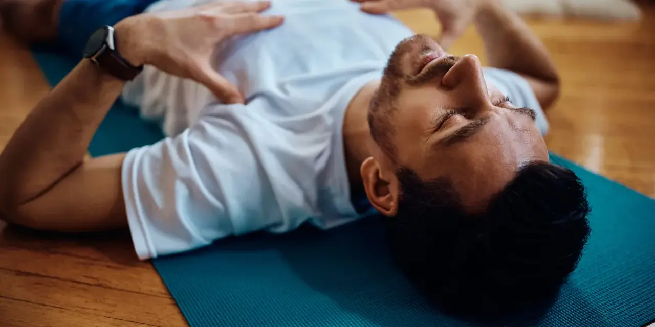 Uomo sdraiato su tappetino blu, mani sul petto, occhi chiusi. Pratica il training autogeno per gestire l'ansia.