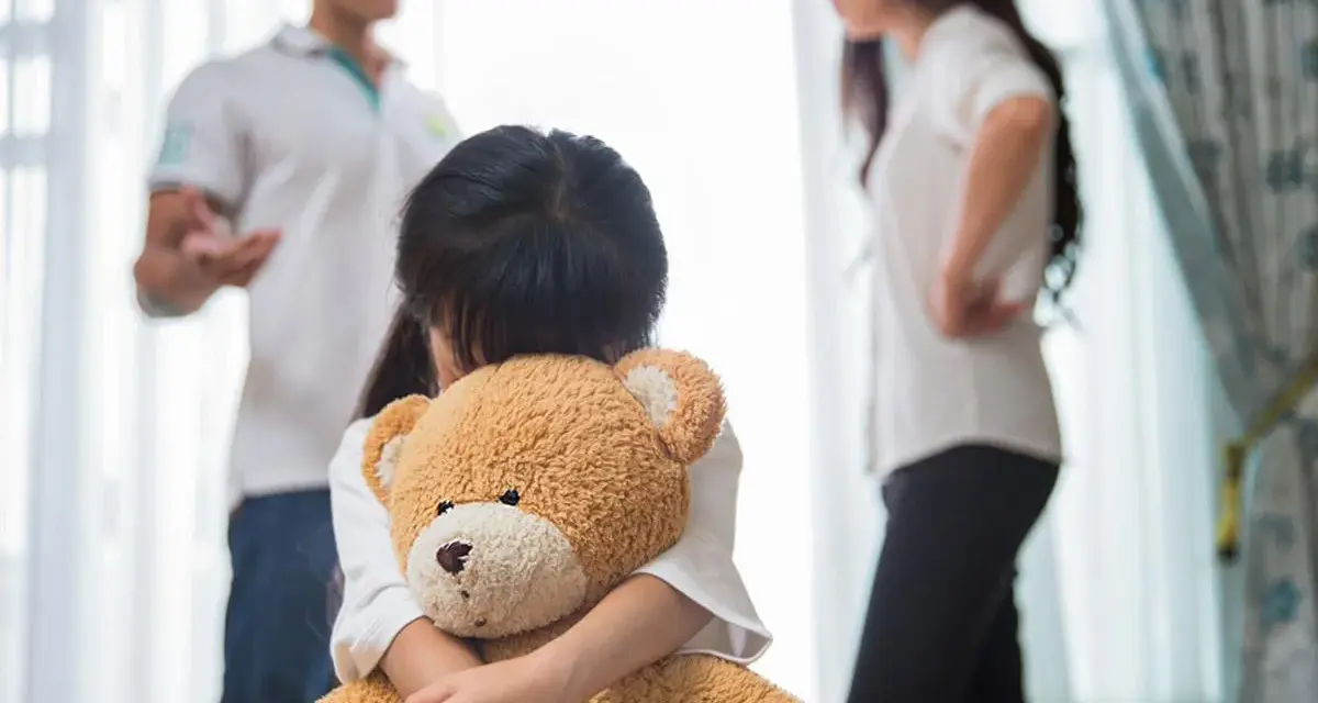 Bambina abbraccia orsacchiotto, mentre i genitori discutono. Avere coraggio di separarsi è difficile per tutti.