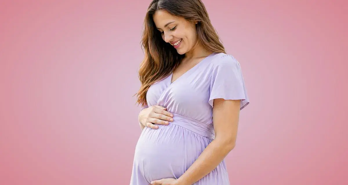 Donna incinta sorride, accarezzando il pancione. Importante assumere acido folico prima o dopo i pasti per la salute del bambino.