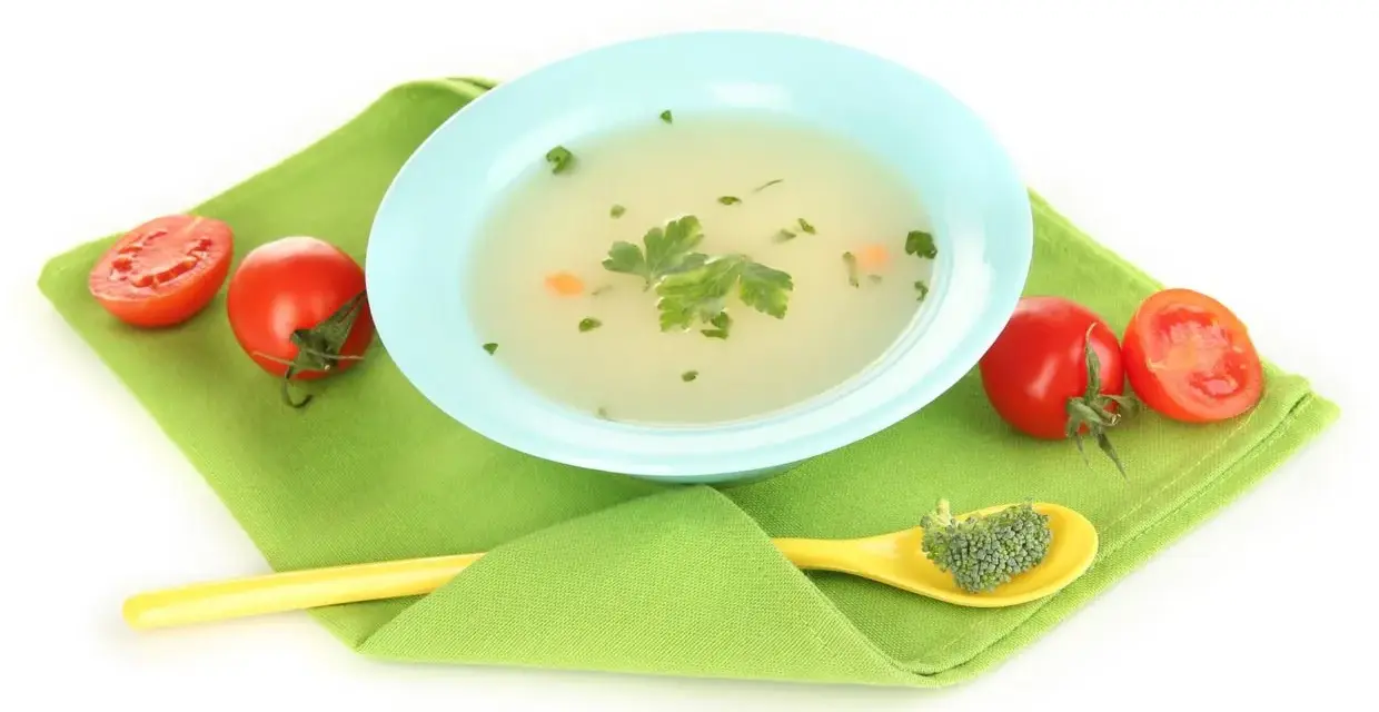 Brodo vegetale svezzamento: una ciotola azzurra con brodo, pomodorini e un cucchiaino giallo con broccoli.