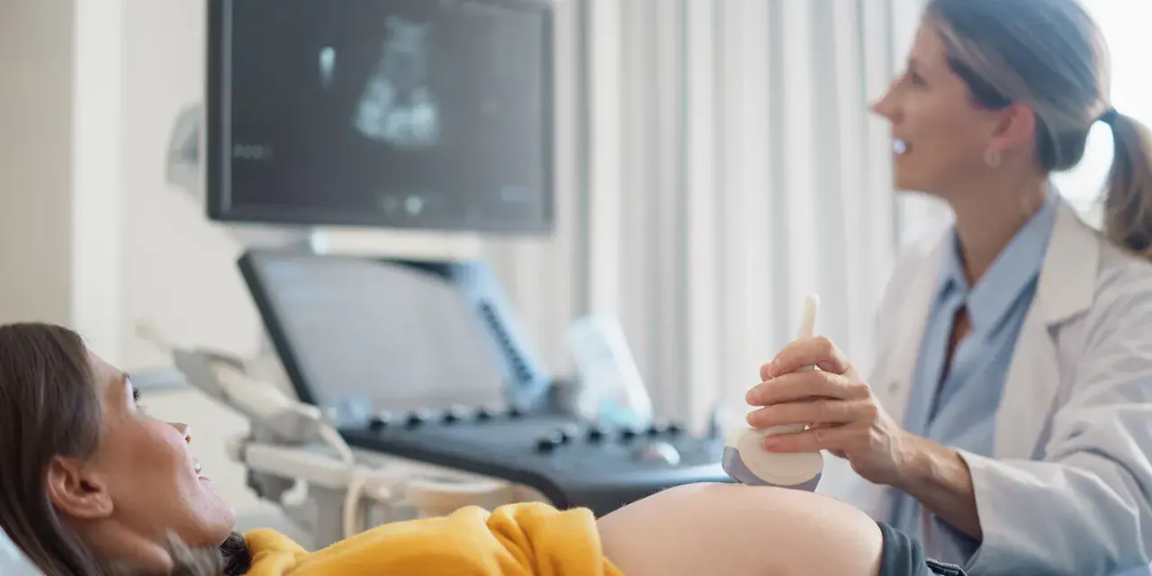 Ecografia di una donna incinta, con il medico che usa la sonda per sentire il battito del feto. L'emozione sul volto della futura mamma è palpabile.