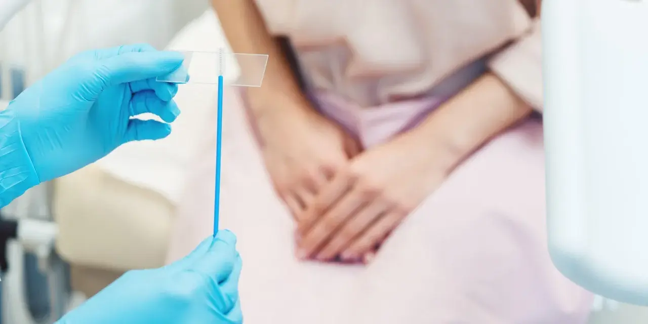 Mani con guanti blu tengono uno strumento per il pap test. La paziente, in attesa, è seduta.