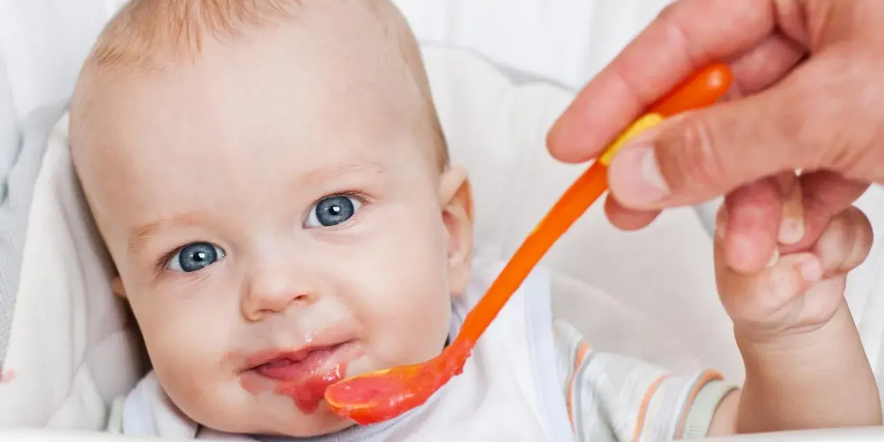 Bambino con occhi azzurri riceve la sua prima pappa svezzamento da un cucchiaino arancione.