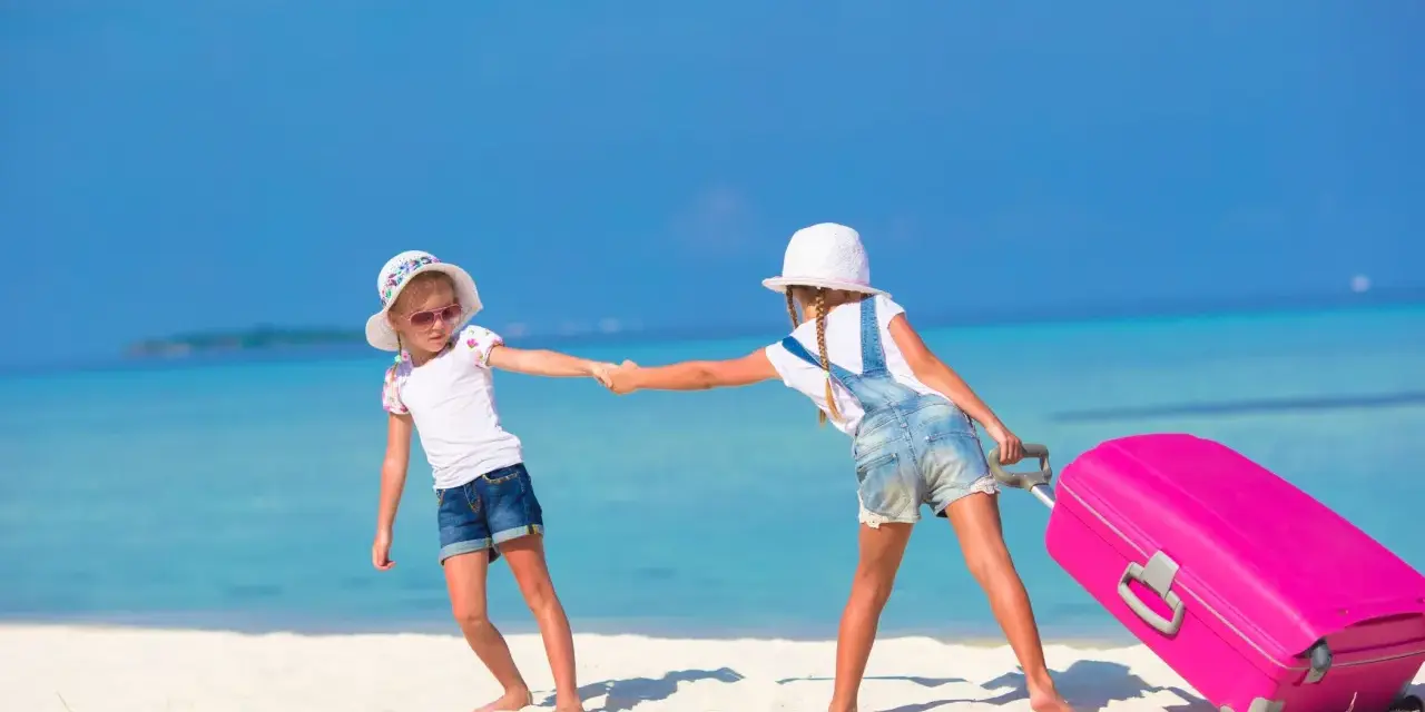 Due bambine con valigia rosa su spiaggia tropicale: idee per cosa fare a Ferragosto con i bambini.
