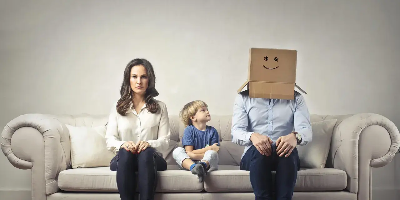 Famiglia seduta sul divano. La madre è triste, il bambino guarda il padre con la testa coperta da un cartone con un sorriso disegnato. Sembra che mio marito stia con me solo per i figli.