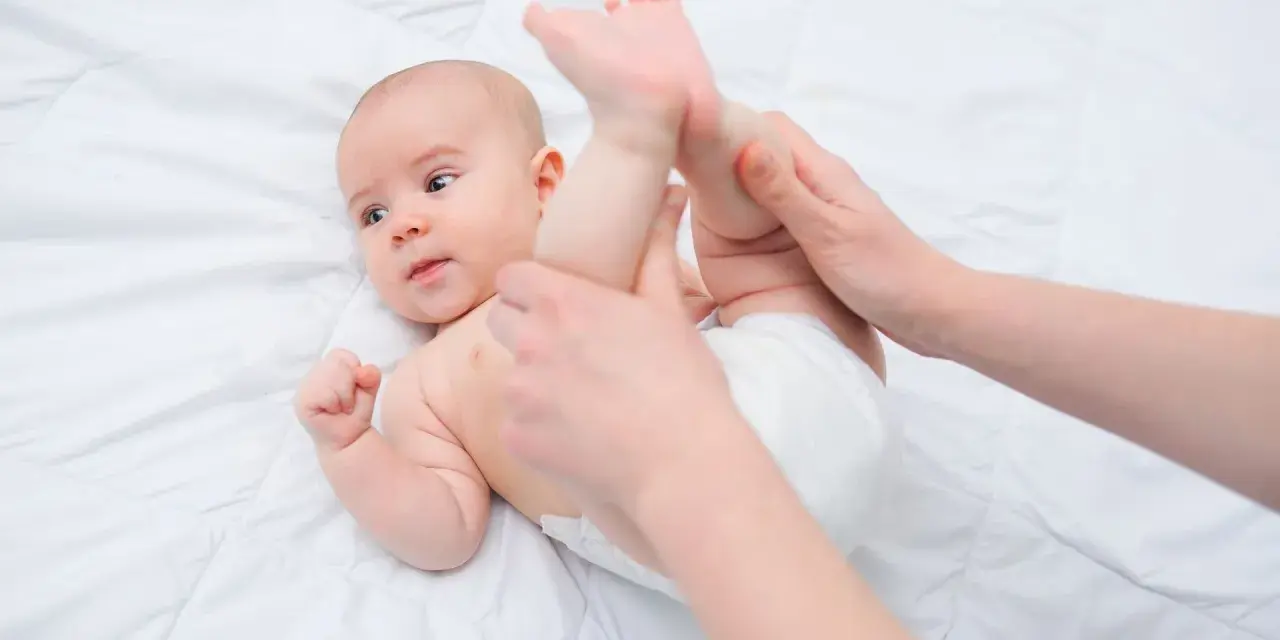 Mani che aiutano un neonato a liberarsi dall'aria nella pancia, con le gambine sollevate delicatamente.
