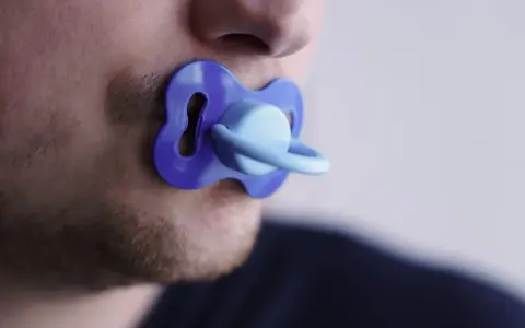 Un uomo con barba corta ha un ciuccio blu in bocca, un'immagine che evoca adulti che si comportano da bambini.