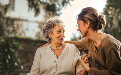 Una donna anziana e una giovane donna si guardano con affetto. La giovane donna ha una mano sulla spalla dell'anziana, un gesto che suggerisce cura e supporto, forse per superare le conseguenze di un cattivo rapporto con la madre.