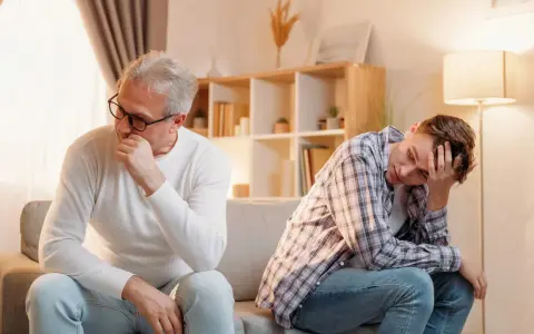 Padre e figlio seduti sul divano, distanti e pensierosi. Crescere in una famiglia dove si litiga sempre lascia segni profondi.