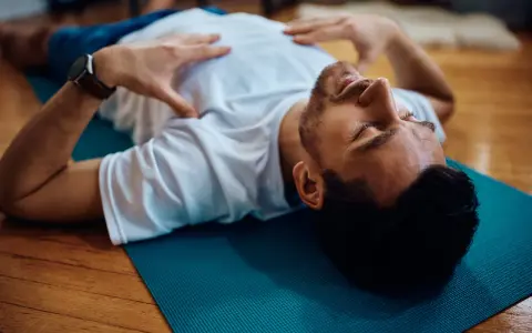 Uomo sdraiato su tappetino blu, mani sul petto, occhi chiusi. Pratica il training autogeno per gestire l'ansia.