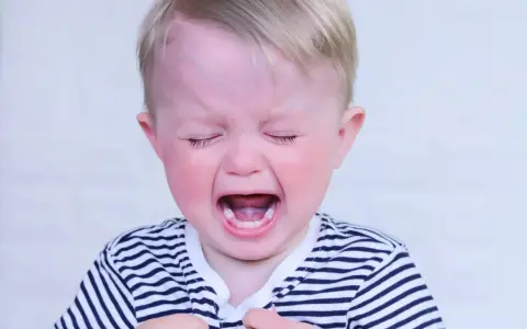 Bambino di 2 anni con capelli biondi, occhi chiusi e bocca spalancata in un pianto disperato, tipico dei capricci continui. Indossa una maglietta a righe.