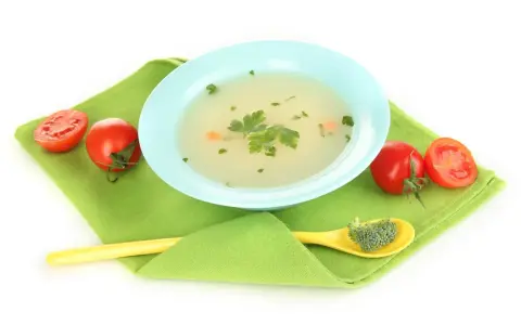 Brodo vegetale svezzamento: una ciotola azzurra con brodo, pomodorini e un cucchiaino giallo con broccoli.