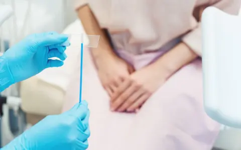 Mani con guanti blu tengono uno strumento per il pap test. La paziente, in attesa, è seduta.
