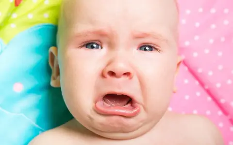 Un neonato piange dopo poppata, con gli occhi azzurri spalancati e la bocca aperta in un lamento.