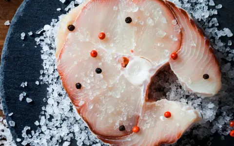 Raw swordfish steak with salt and peppercorns on a dark slate.