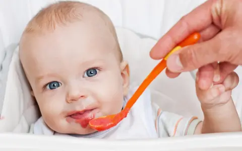 Bambino con occhi azzurri riceve la sua prima pappa svezzamento da un cucchiaino arancione.