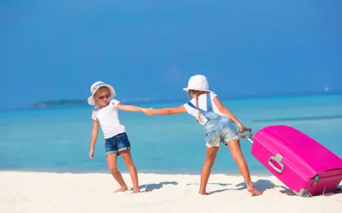 Due bambine con valigia rosa su spiaggia tropicale: idee per cosa fare a Ferragosto con i bambini.