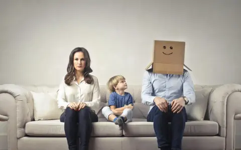 Famiglia seduta sul divano. La madre è triste, il bambino guarda il padre con la testa coperta da un cartone con un sorriso disegnato. Sembra che mio marito stia con me solo per i figli.
