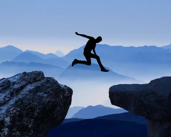 Silhouette di una persona che salta tra due rocce, con montagne blu sullo sfondo. Un viaggio per perdersi per ritrovarsi, un salto nel vuoto per una coppia.