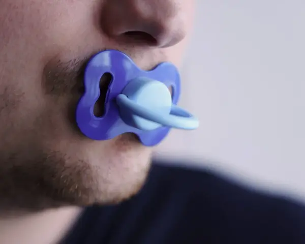 Un uomo con barba corta ha un ciuccio blu in bocca, un'immagine che evoca adulti che si comportano da bambini.