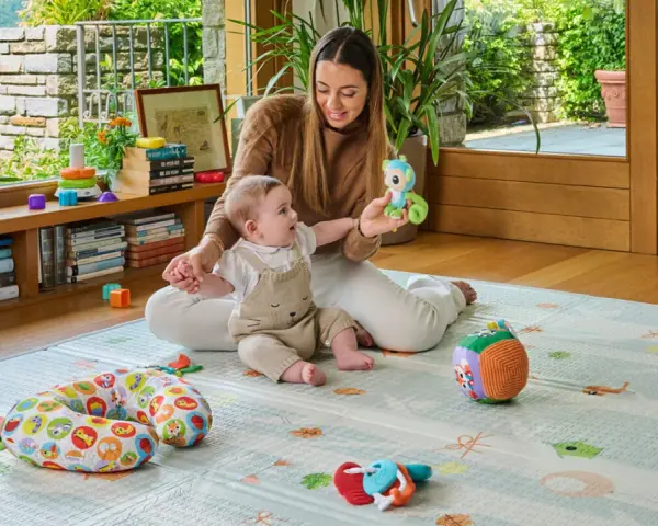 Mamma gioca con un neonato di 6 mesi che non sta seduto, su un tappeto gioco con tanti giocattoli colorati.
