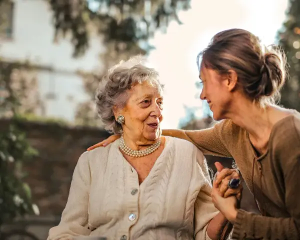 Una donna anziana e una giovane donna si guardano con affetto. La giovane donna ha una mano sulla spalla dell'anziana, un gesto che suggerisce cura e supporto, forse per superare le conseguenze di un cattivo rapporto con la madre.