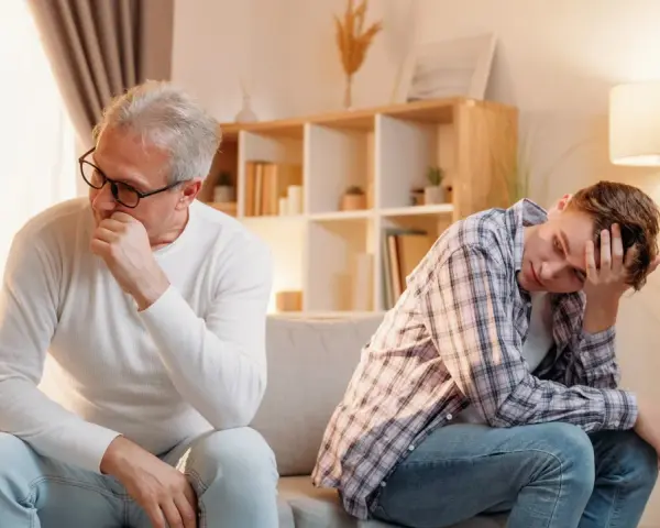 Padre e figlio seduti sul divano, distanti e pensierosi. Crescere in una famiglia dove si litiga sempre lascia segni profondi.