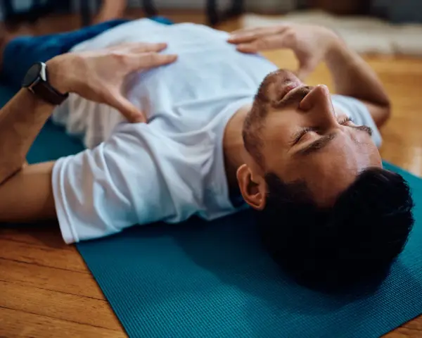 Uomo sdraiato su tappetino blu, mani sul petto, occhi chiusi. Pratica il training autogeno per gestire l'ansia.