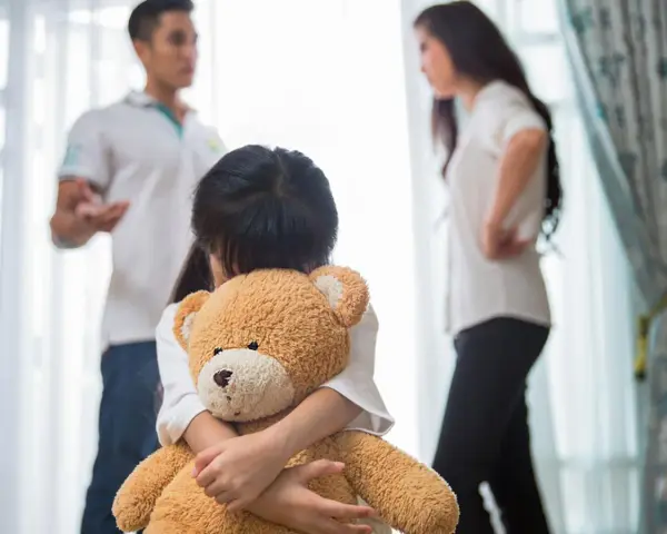 Bambina abbraccia orsacchiotto, mentre i genitori discutono. Avere coraggio di separarsi è difficile per tutti.
