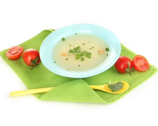 Brodo vegetale svezzamento: una ciotola azzurra con brodo, pomodorini e un cucchiaino giallo con broccoli.