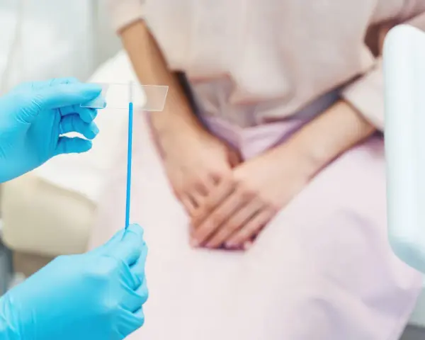 Mani con guanti blu tengono uno strumento per il pap test. La paziente, in attesa, è seduta.