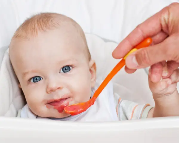 Bambino con occhi azzurri riceve la sua prima pappa svezzamento da un cucchiaino arancione.