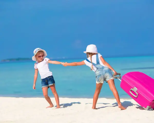 Due bambine con valigia rosa su spiaggia tropicale: idee per cosa fare a Ferragosto con i bambini.