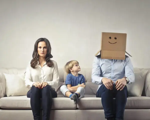 Famiglia seduta sul divano. La madre è triste, il bambino guarda il padre con la testa coperta da un cartone con un sorriso disegnato. Sembra che mio marito stia con me solo per i figli.