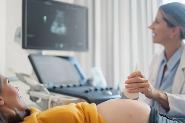 Ecografia di una donna incinta, con il medico che usa la sonda per sentire il battito del feto. L'emozione sul volto della futura mamma è palpabile.