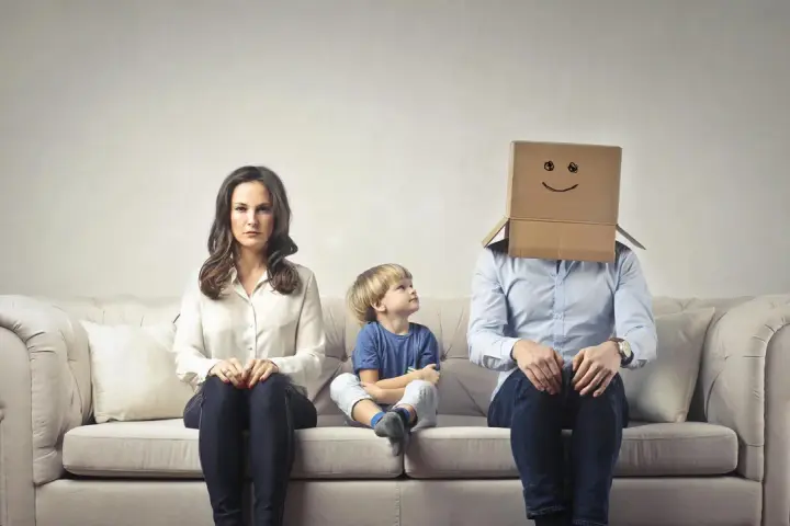 Famiglia seduta sul divano. La madre è triste, il bambino guarda il padre con la testa coperta da un cartone con un sorriso disegnato. Sembra che mio marito stia con me solo per i figli.