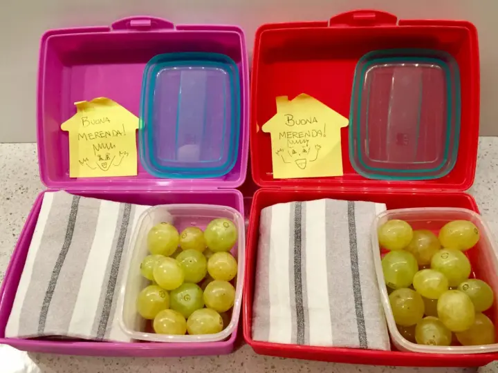 Due lunchbox rosa e rosse pronte per le merende per bambini, con uva e un bigliettino "Buona Merenda!".