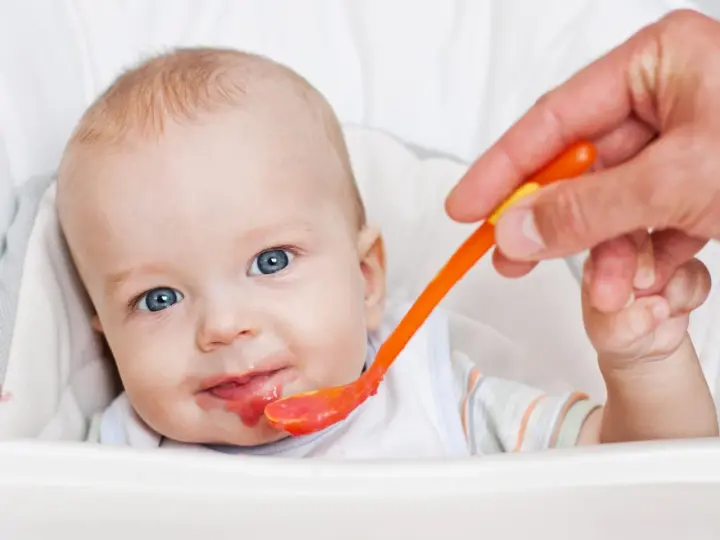 Bambino con occhi azzurri riceve la sua prima pappa svezzamento da un cucchiaino arancione.