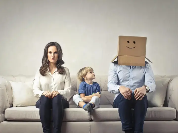 Famiglia seduta sul divano. La madre è triste, il bambino guarda il padre con la testa coperta da un cartone con un sorriso disegnato. Sembra che mio marito stia con me solo per i figli.