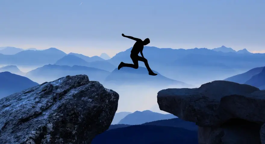 Silhouette di una persona che salta tra due rocce, con montagne blu sullo sfondo. Un viaggio per perdersi per ritrovarsi, un salto nel vuoto per una coppia.